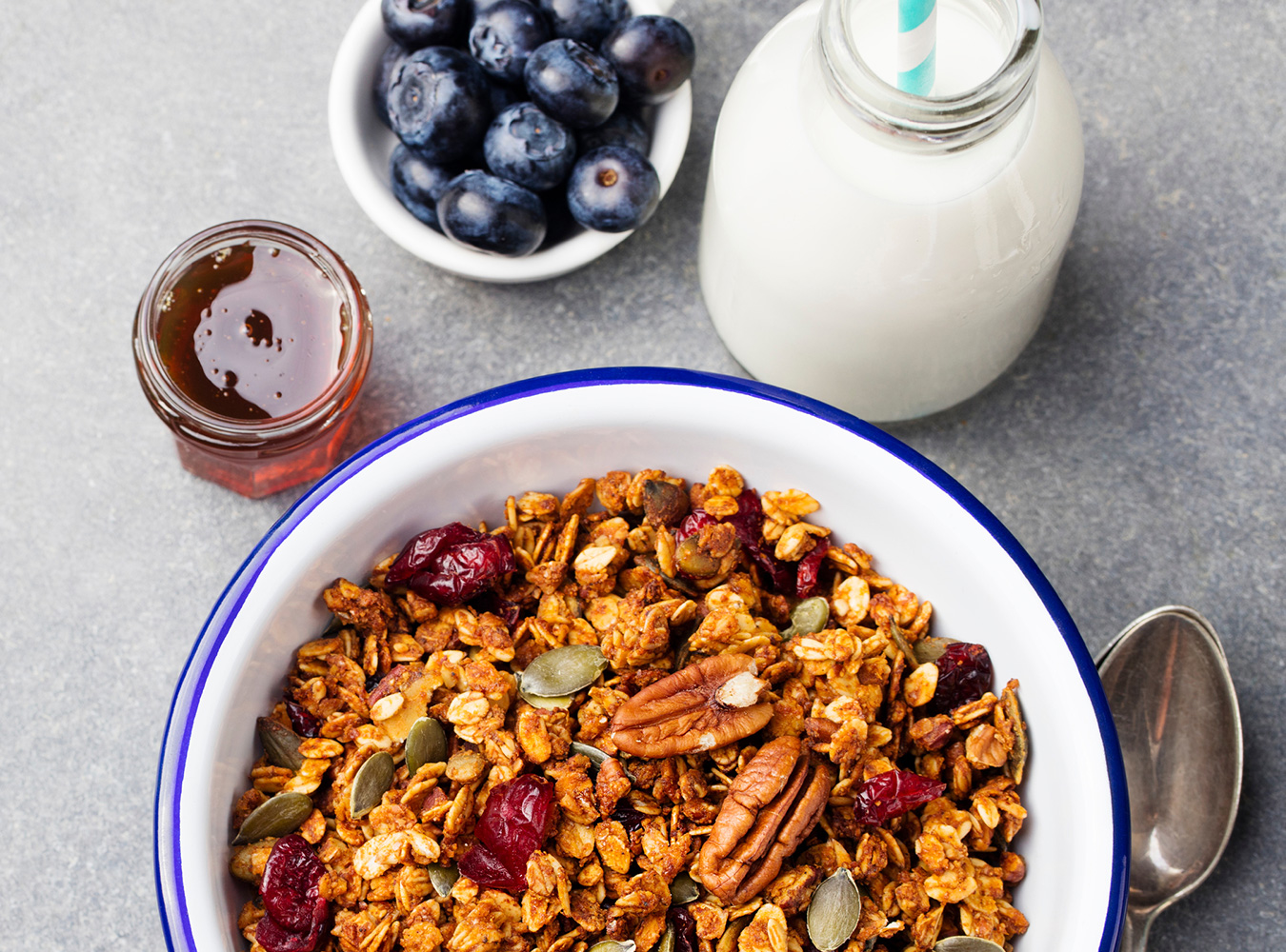 A bowl of oats with nuts, seeds and dried fruit next to blueberries, honey and milk