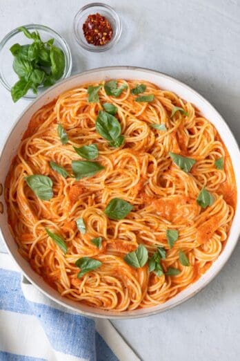 Pasta tossed in a creamy roasted red pepper sauce garnished with freshly torn basil leaves and sides of basil and red pepper flakes next to skillet.