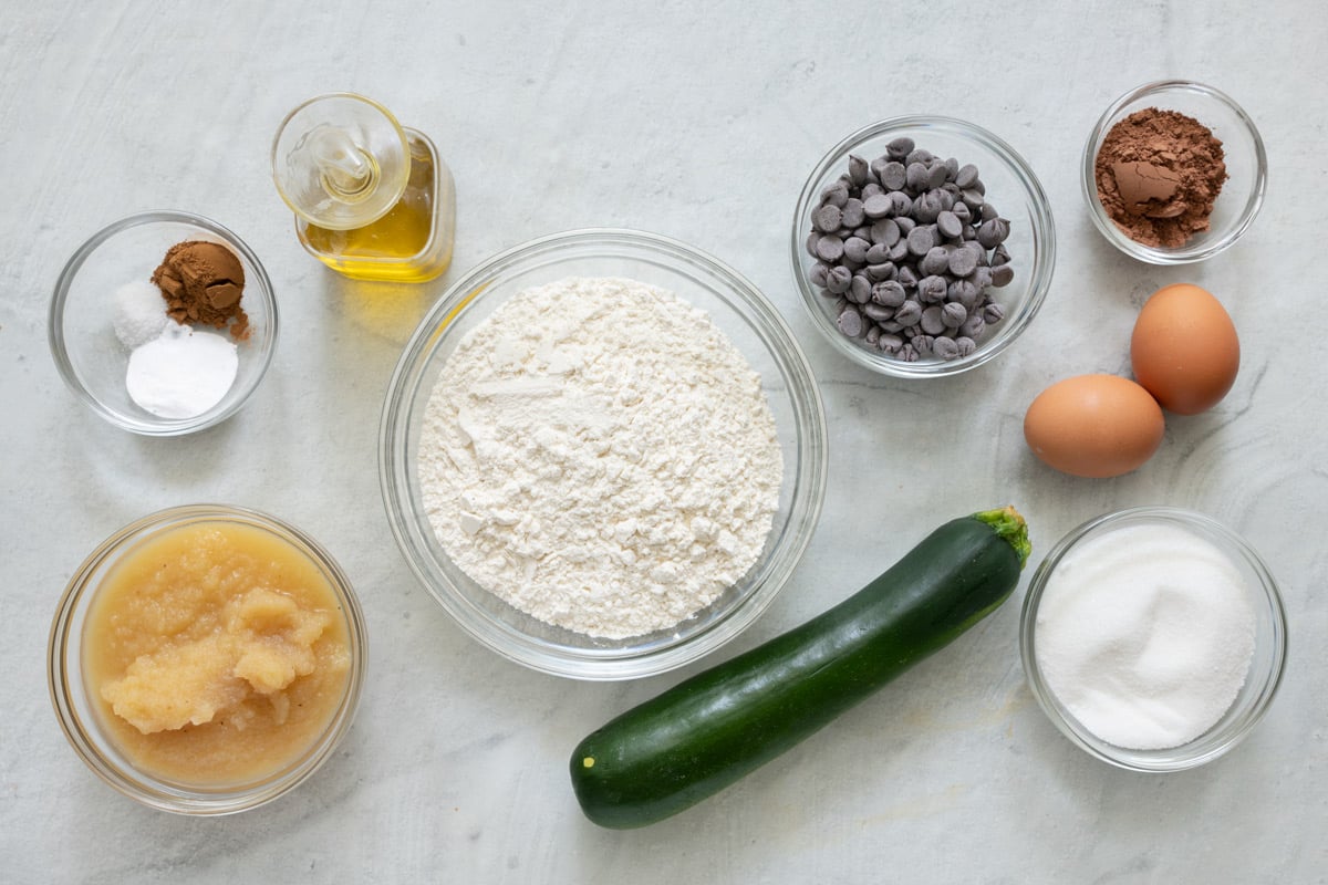Ingredients for bread recipe before being prepped: spices, applesauce, oil, flour, zucchini, chocolate chips, cocoa powder, 2 eggs, and sugar.