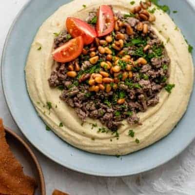 Blue plate with hummus, ground beef, pine nuts, parsley and tomatoes, with side of fried pita chips