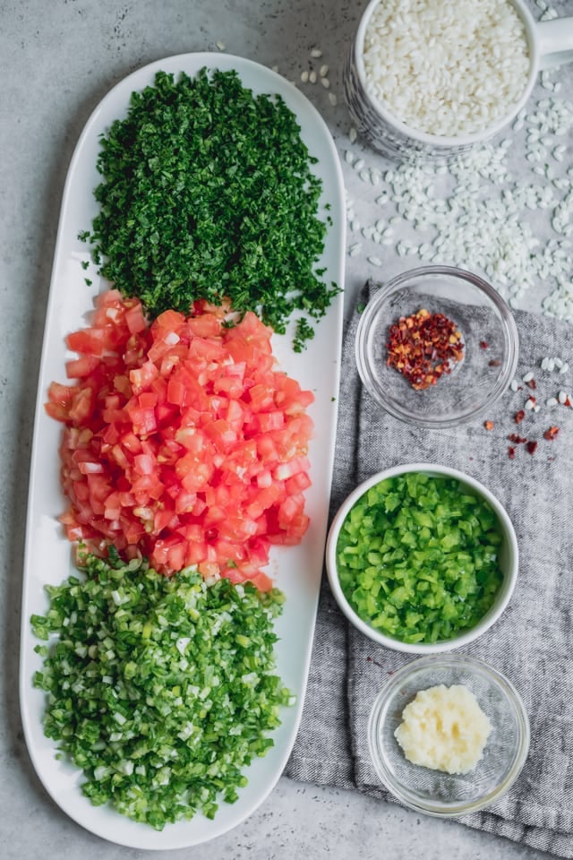 Ingredients for Vegetarian Stuffed Grape Leaves - parsley, tomatoes and green onions on a long white plate. Additionally, there is a cup of short grain rice, green peppers, garlic and crushed red pepper.