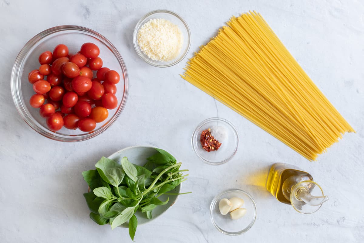 Ingredients for pasta recipe: bowl of cherry tomatoes, fresh basil, Parmesan cheese, spices, garlic cloves, olive oil, and dry noodles.