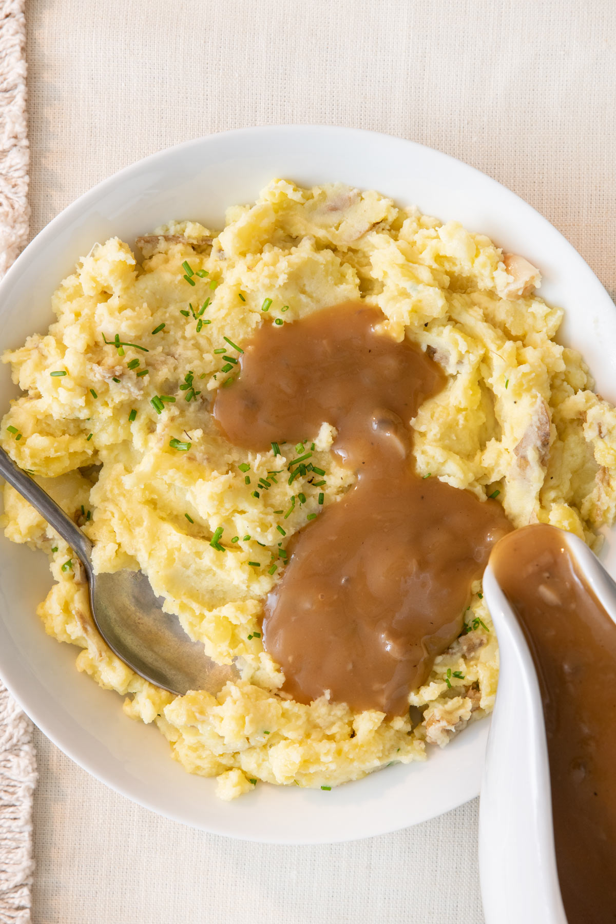 Gravy being poured over a serving dish full of mashed potatoes garnished with chopped chives and a serving spoon inside bowl.