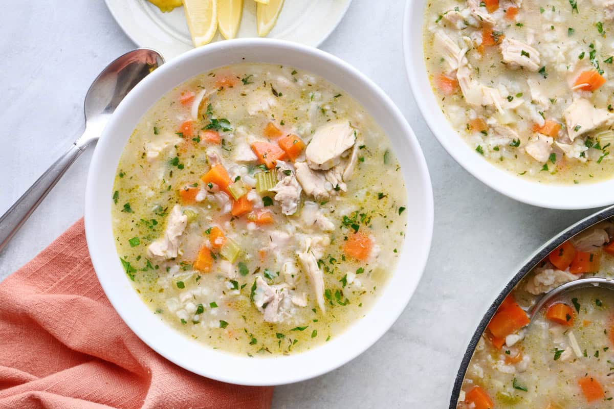 Bowl of lemon chicken rice soup, with a a second bowl and pot nearby and extra lemon wedges on a plate