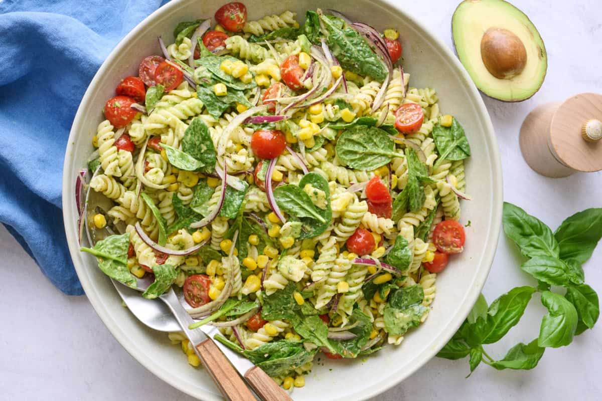 Avocado pasta salad in a serving bowl with serving set dipped inside, halved avocado nearby.