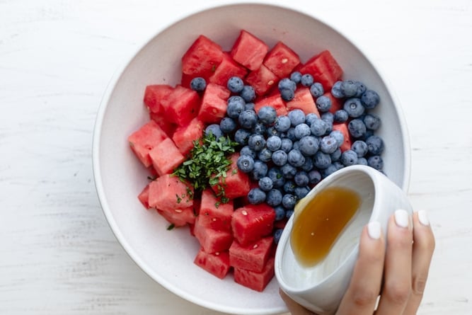 Large white bowl of cubed watermelon pieces with blueberries and mint; pouring honey and lemon mixture over