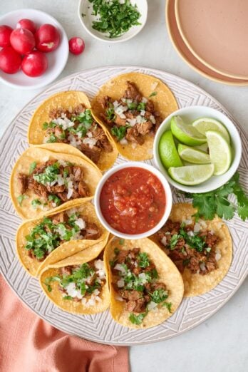 Homemade Mexican Street Tacos on a round platter with a small dish of salsa, garnished with fresh chopped cilantro and onions.