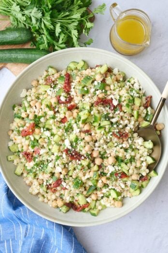 Couscous salad in a wide shallow bowl garnished with extra feta, serving spoon dipped inside with small jar of extra dressing nearby.