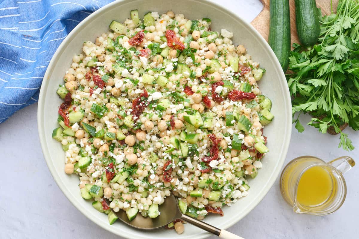 Couscous salad in a wide shallow bowl garnished with extra feta, serving spoon dipped inside with small jar of extra dressing nearby.