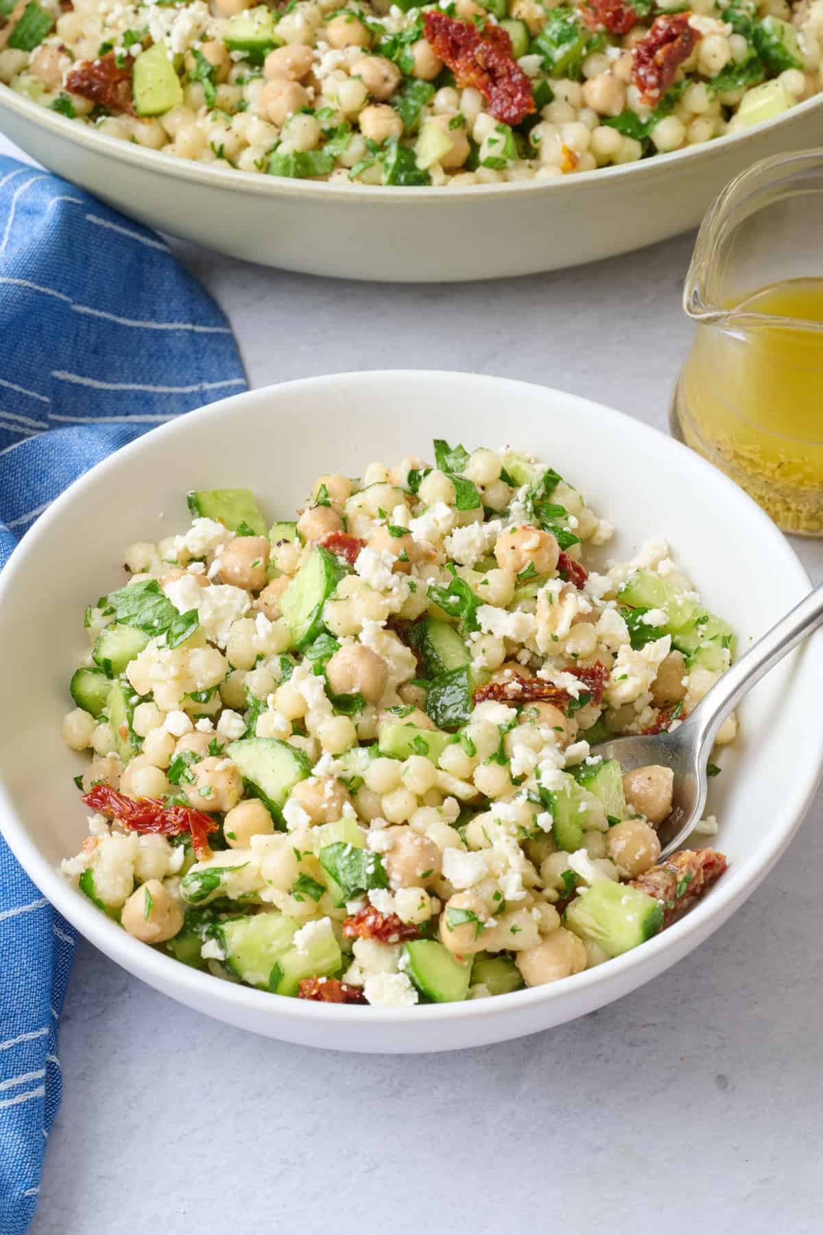 A serving of couscous salad in a small bowl with serving bowl and dressing jar nearby.