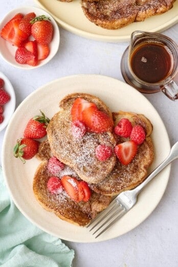 Vegan French Toast on a plate topped with strawberries, with more toppings and another plate of the french toast surrounding.