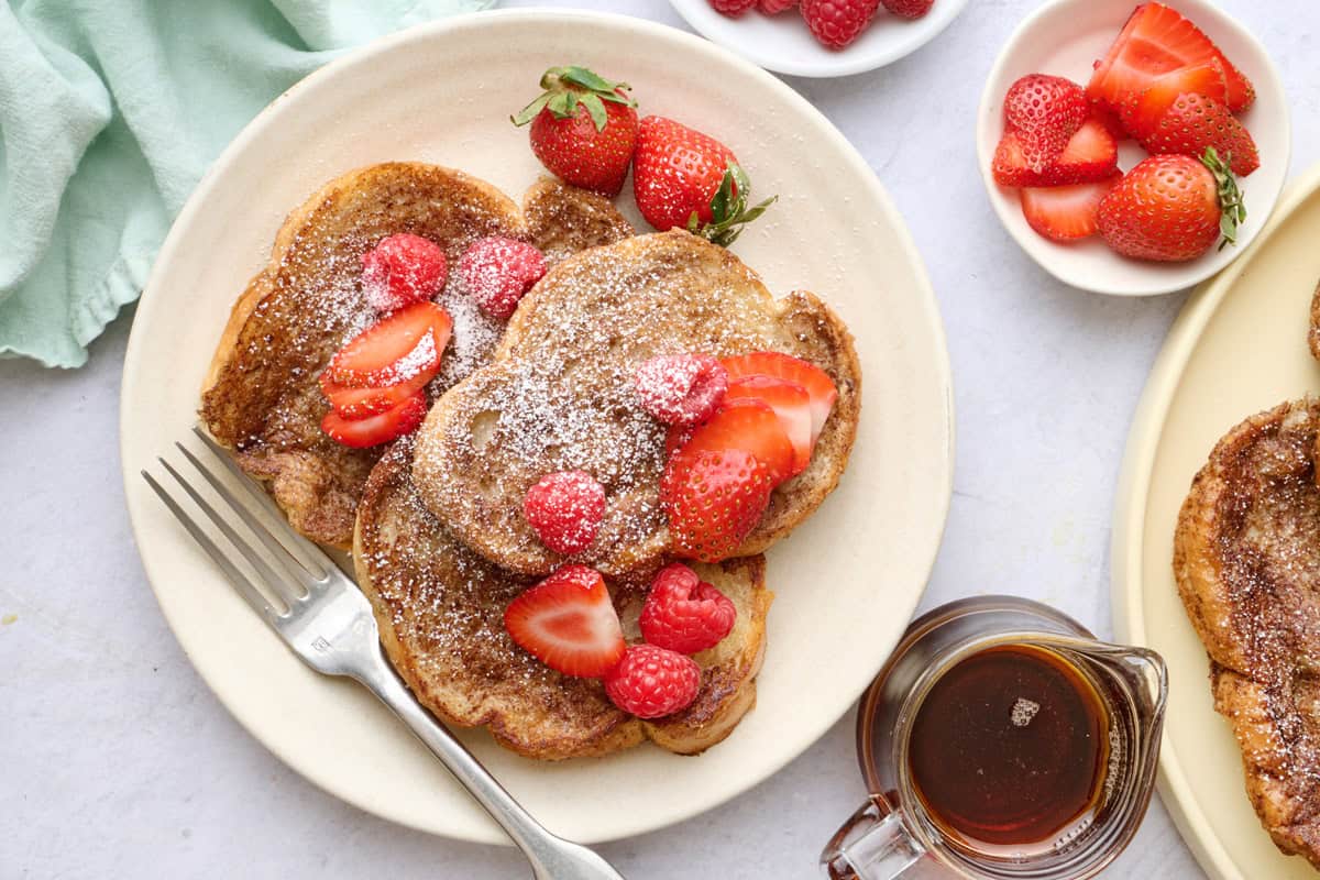 Vegan French Toast on a plate topped with strawberries, with more toppings and another plate of the french toast surrounding.  