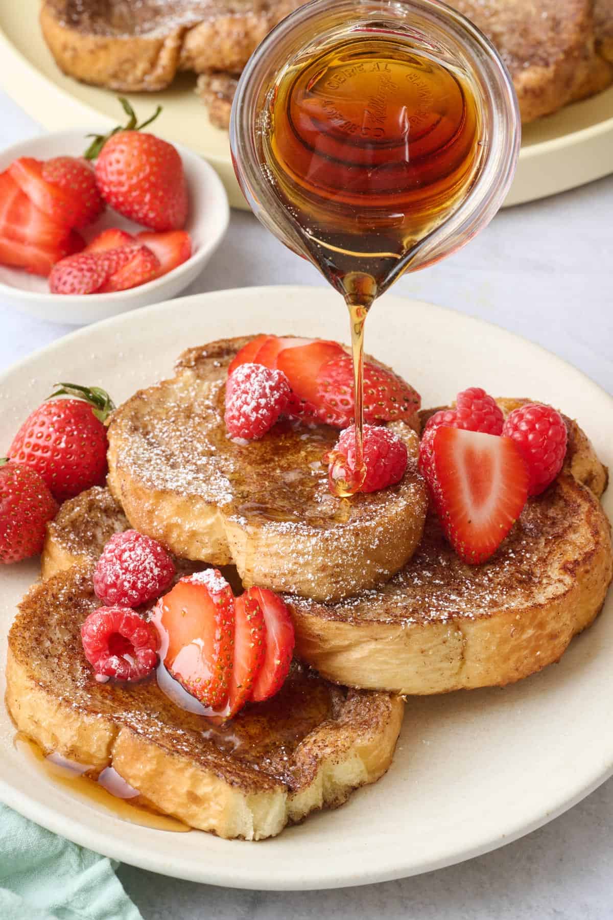 Three pieces of Vegan French Toast stacked on a plate, topped with strawberries, with syrup being poured on top.