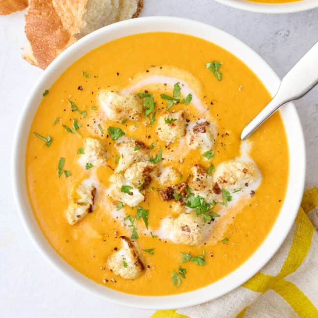 Two bowls of soup with a little extra swirl of coconut milk and some roasted cauliflower spooned on top, garnished with cilantro, crust bread nearby.