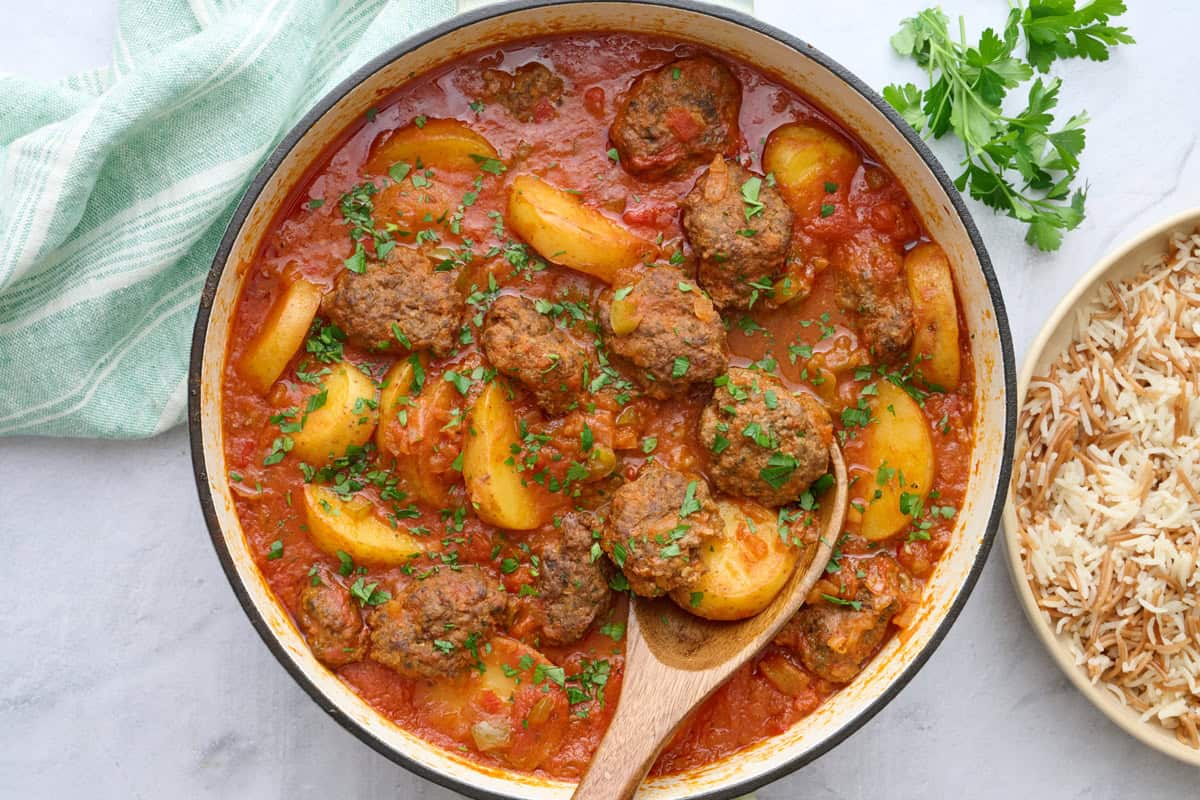 Kafta Potato Stew in the pot garnished lightly with chopped parsley, serving spoon, small dish of chopped parsley, and serving dish of Lebanese rice nearby.