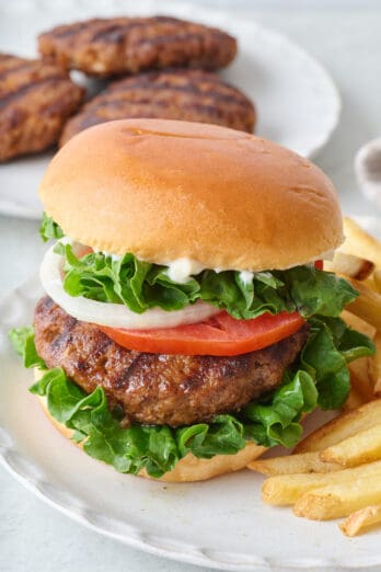 Homemade hamburger assembled with bun and toppings with a side of fries and more grilled patties in back.