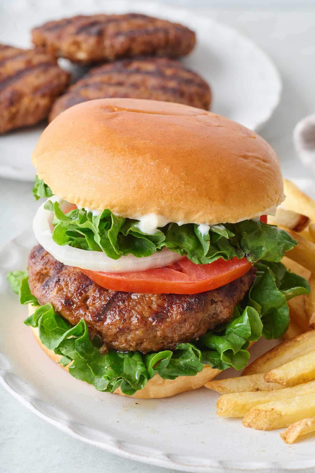Homemade hamburger assembled with bun and toppings with a side of fries and more grilled patties in back.