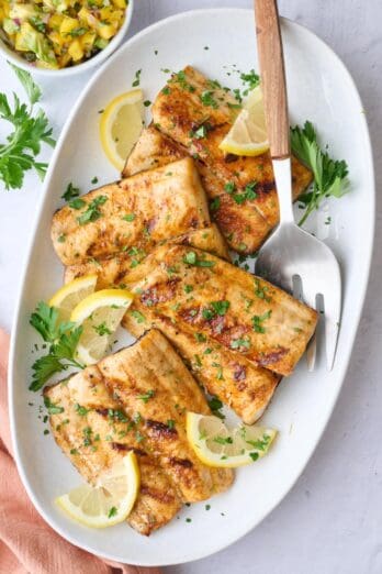 Grilled mahi mahi fillets on a large platter garnished with chopped parsley and lemon wedges, small bowl of mango salsa nearby.
