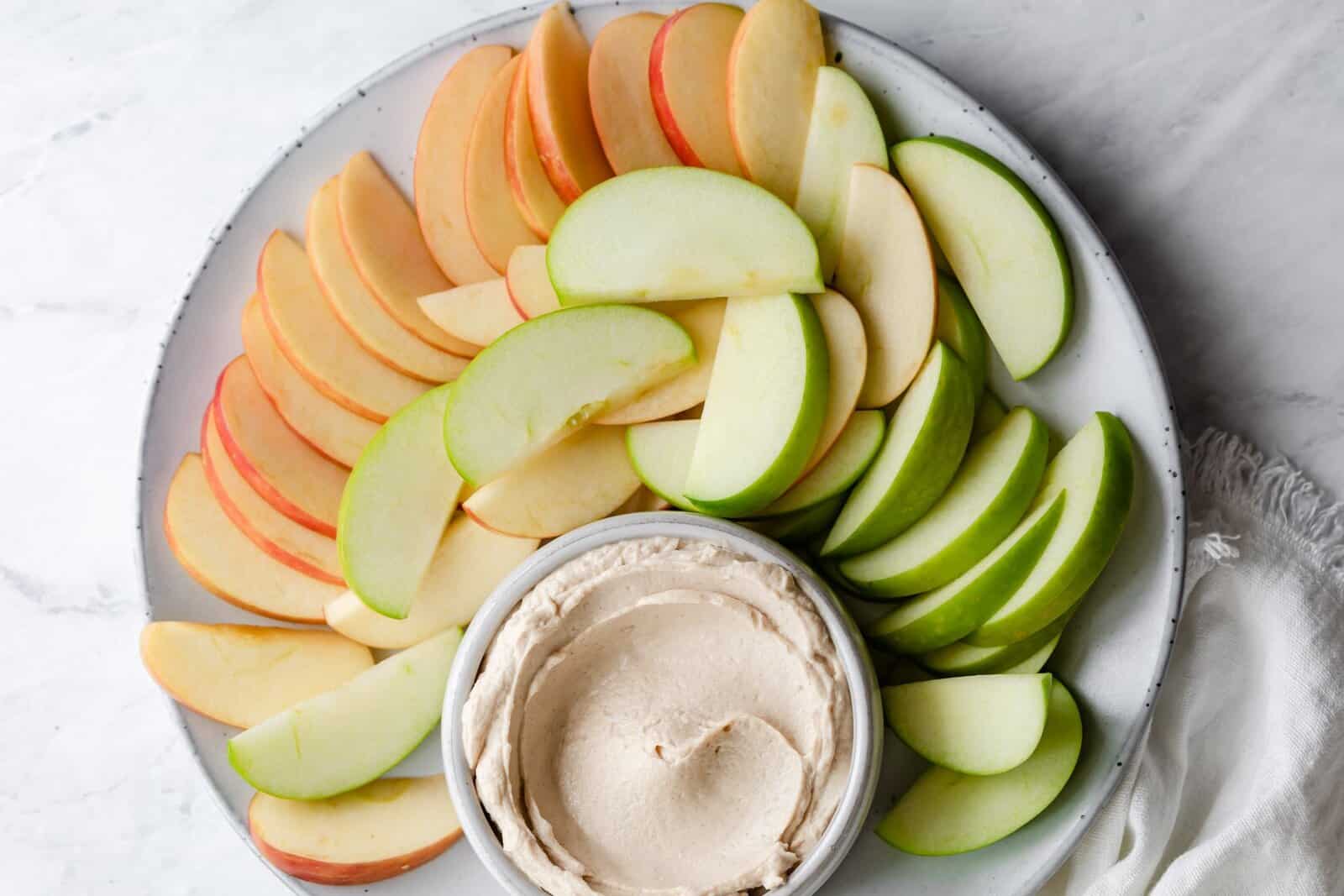 Peanut butter dip in a small bowl that is placed on top of a plate of cut apples. 