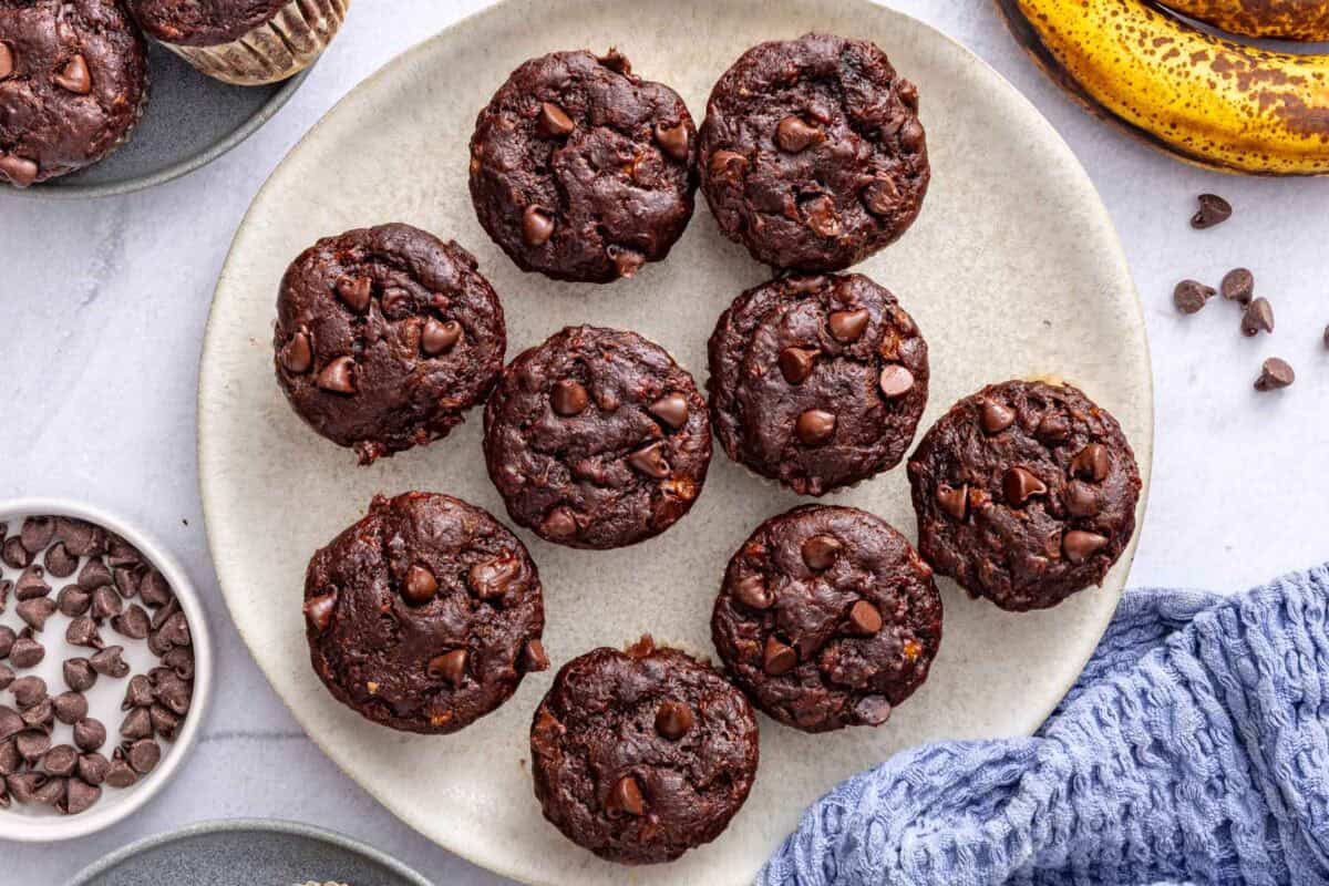 Chocolate banana muffins on a plate with a few on small plates nearby, bananas and small dish of chocolate chips around.