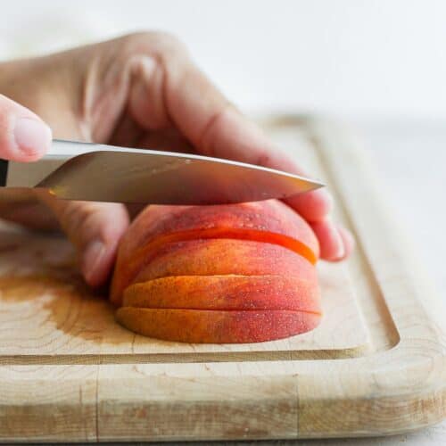 Half of a peach face down on a cutting board with a knife cutting slices.