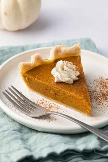 Slice of pumpkin pie on a small plate with whipped cream on top with cinnamon sprinkled on top and a fork sitting on plate.