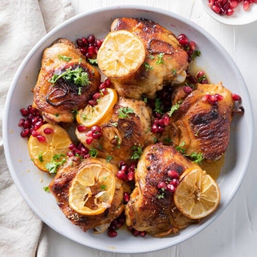 Pomegranate chicken thighs in a serving bowl with shallots, lemons, and garnish.