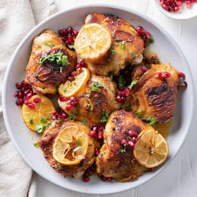 Pomegranate chicken thighs in a serving bowl with shallots, lemons, and garnish.