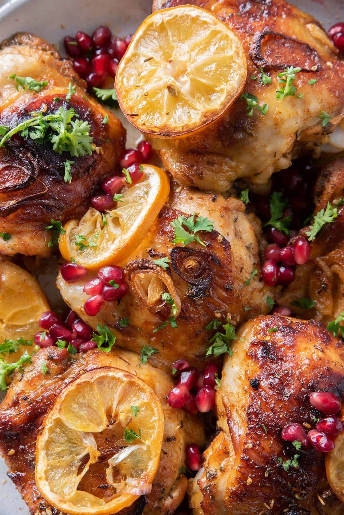 Pomegranate chicken thighs in a serving bowl with shallots, lemons, and garnish.