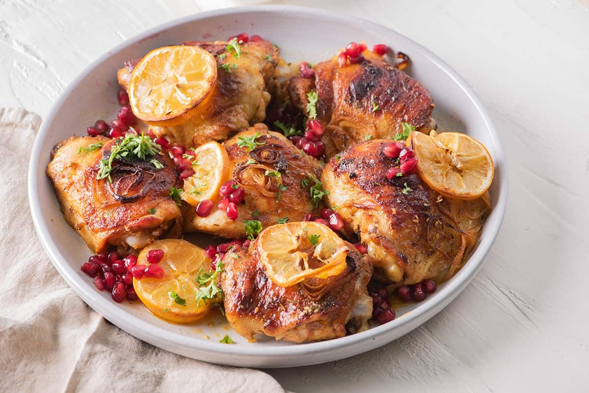 Pomegranate chicken thighs in a serving bowl with shallots, lemons, and garnish.
