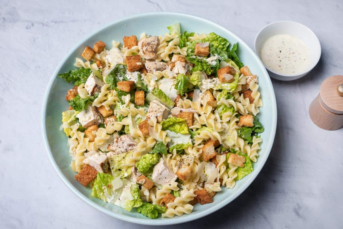 Chicken caesar pasta salad in a bowl with ingredients nearby.