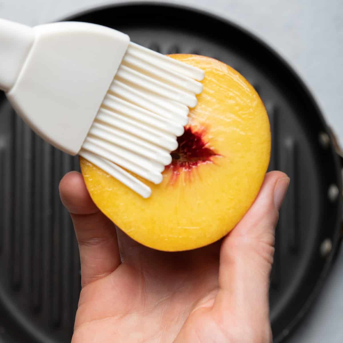 Hand holding half peach brushing melted coconut oil on it over grill pan.