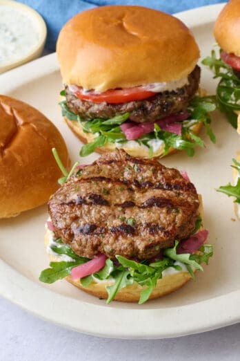 Assembled burgers on a platter topped with yogurt, lid off one burger, and yogurt dish nearby.