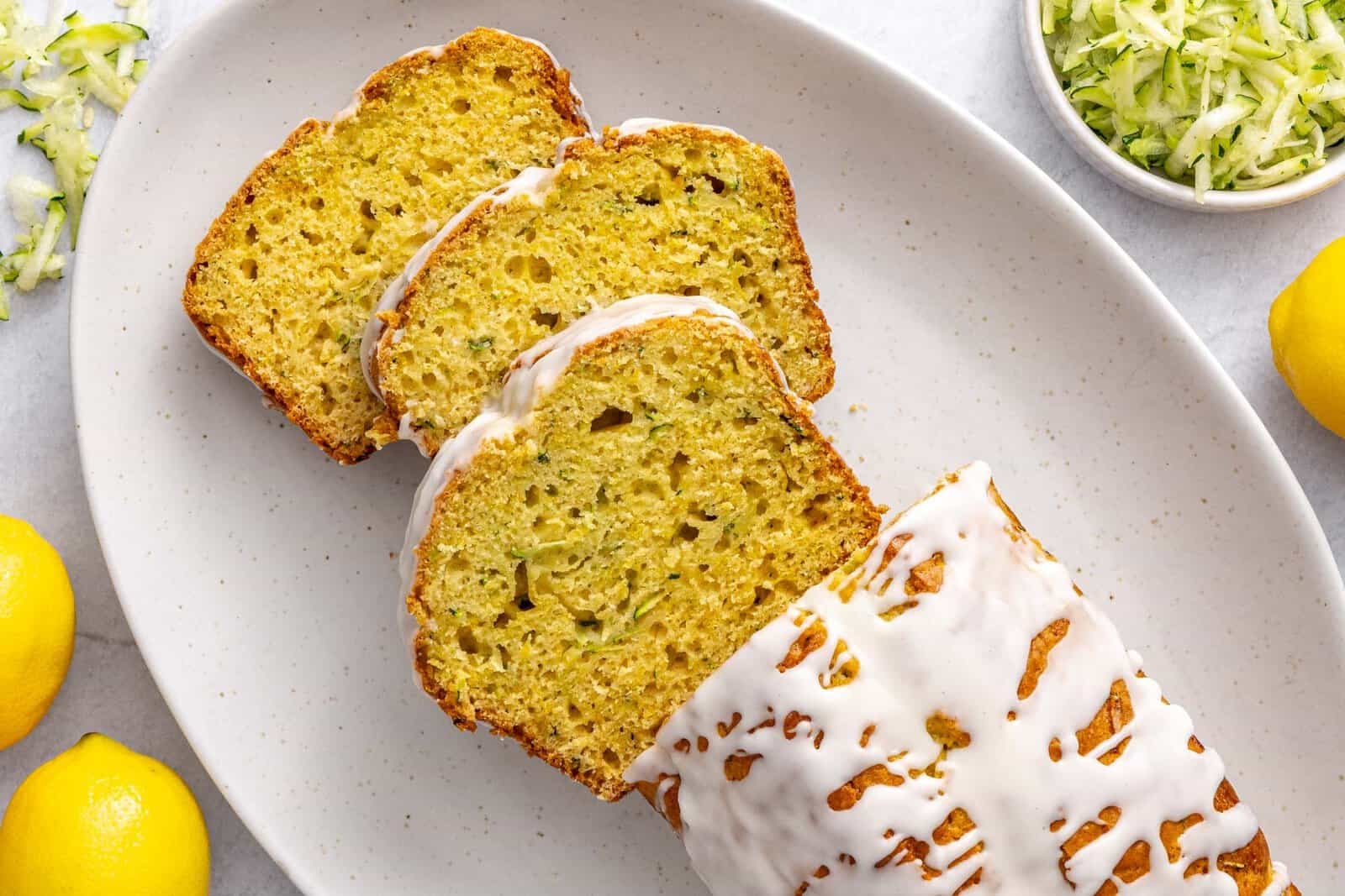 Lemon zucchini loaf on a platter drizzled with icing and garnished lightly with lemon zest. A few thick slices cut and leaning forward.
