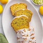 Lemon zucchini loaf on a platter drizzled with icing and garnished lightly with lemon zest. A few thick slices cut and leaning forward.