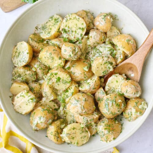 Tahini potato salad in a round serving bowl garnished with extra chopped herbs with lemon wedges in a small dish nearby, serving spoon nearby.