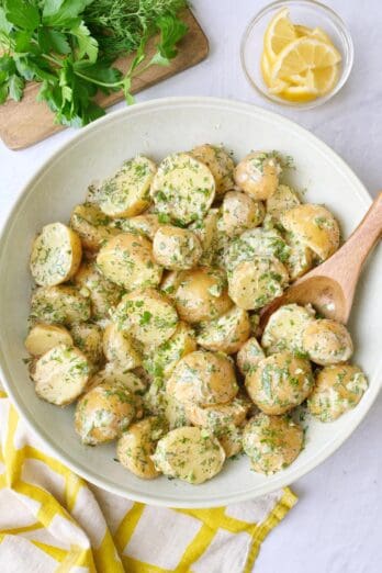 Tahini potato salad in a round serving bowl garnished with extra chopped herbs with lemon wedges in a small dish nearby, serving spoon nearby.
