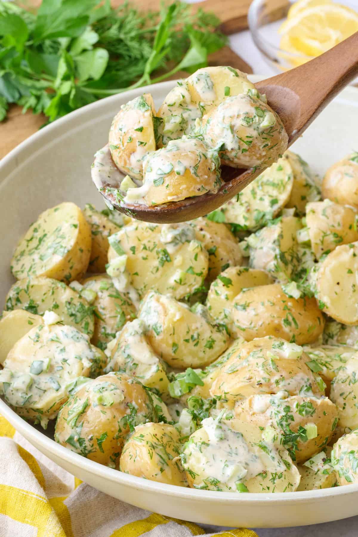 Tahini potato salad in a round serving bowl garnished with extra chopped herbs and a serving spoon holding up some.