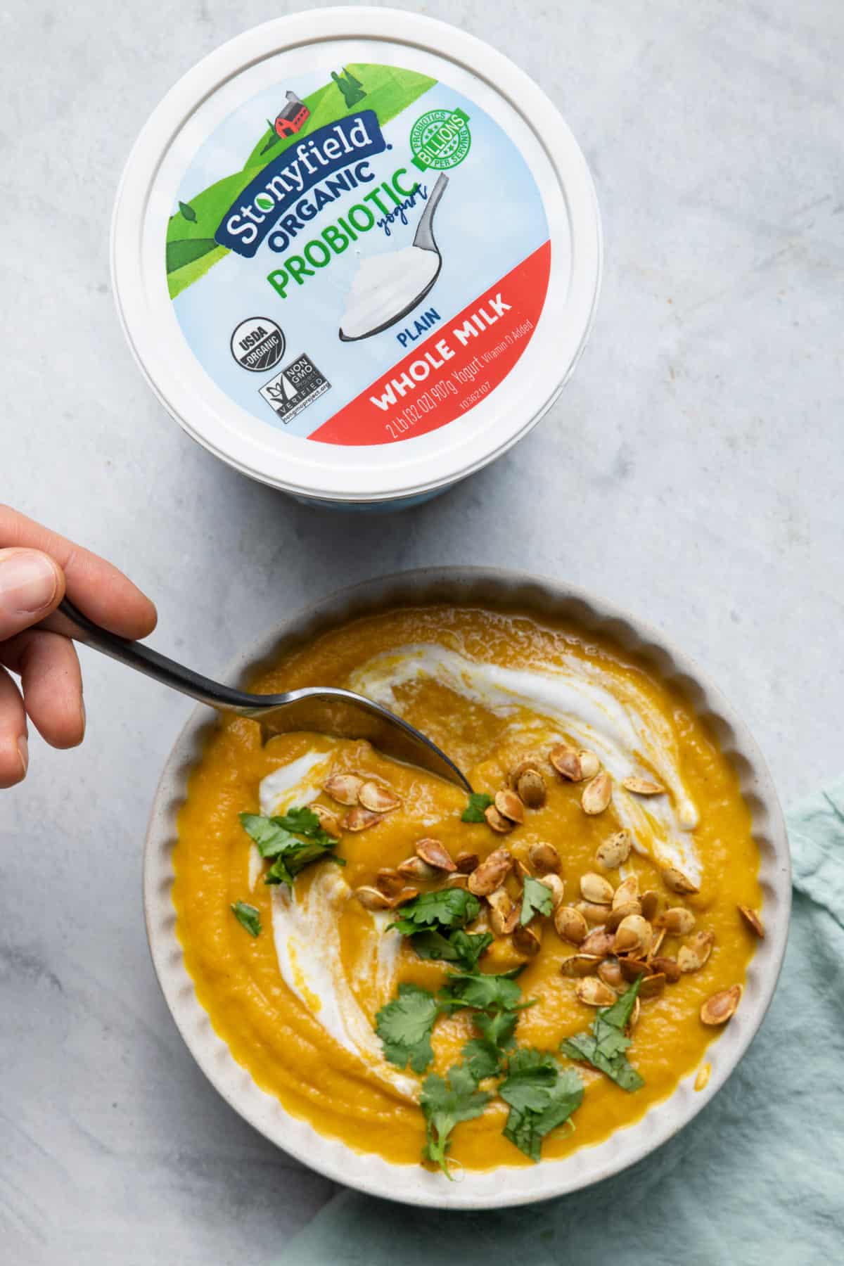 Spoon taking bite out of acorn squash soup with tub of Stonyfield yogurt next to it used to make the soup
