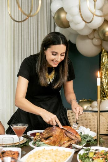 Yumna slicing into the Thanksgiving turkey carving a piece of the breast with fork and knife with the platter surrounded with side dishes and dinnerware.