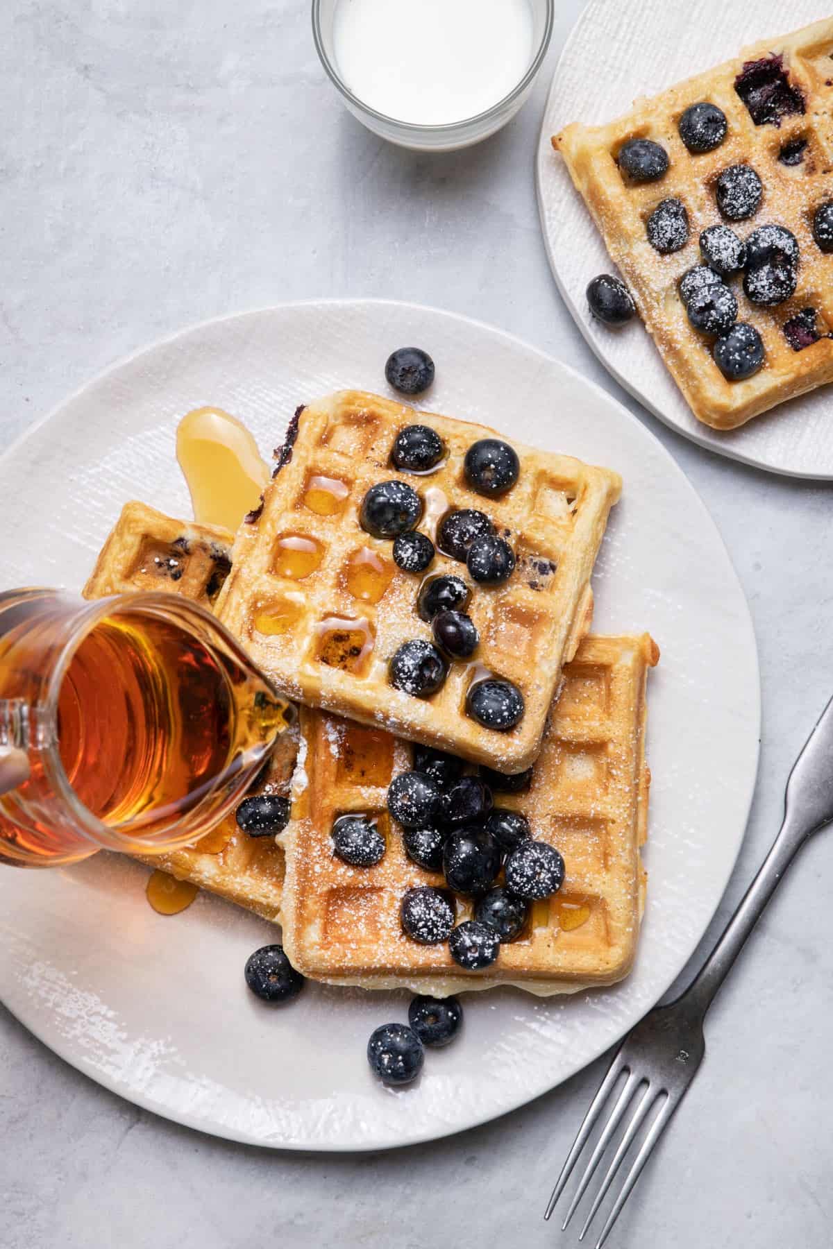 Plate of waffles with blueberries, powdered sugar and maple syrup being poured on top.