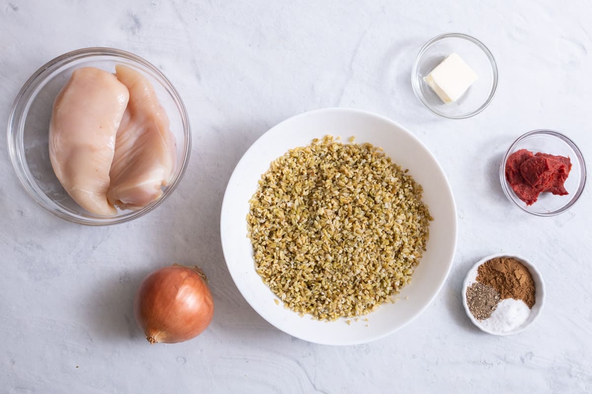 ingredients for recipe with 2 pieces of chicken breast in bowl, bowl of freekeh, whole yellow onion, butter, tomato paste, and spices