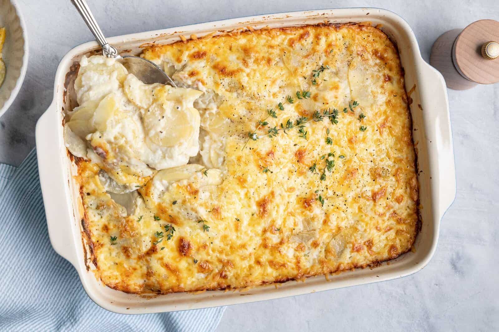 Scalloped potatoes after done baking garnished with fresh thyme leaves and black pepper and a serving spoon removing a scoop.