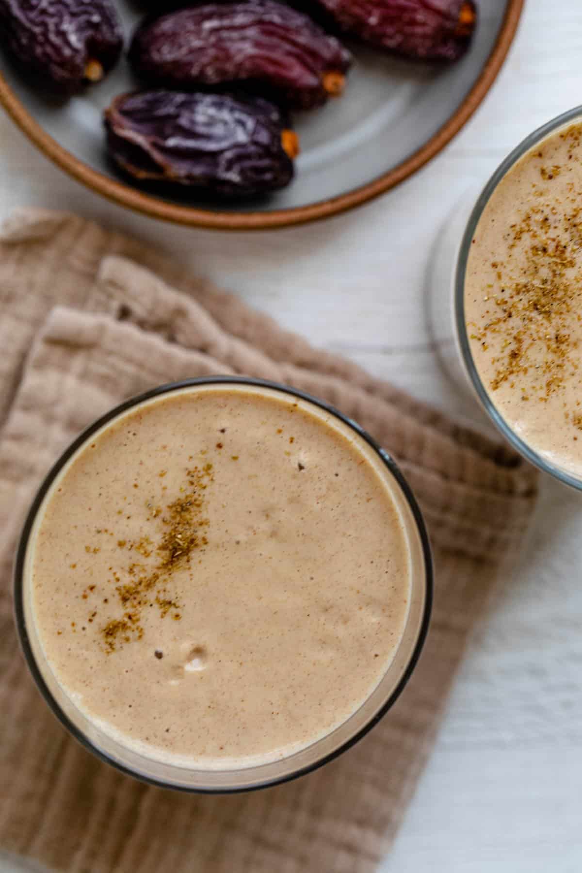 Overhead shot of smoothie recipe with cardamom sprinkled on top and a bowl of dates in a bowl slightly of shot.