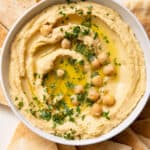 Overhead shot of recipe topped with oil, chickpeas, and parsley in a bowl served with pita chips on large plate.