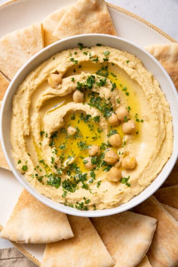 Overhead shot of recipe topped with oil, chickpeas, and parsley in a bowl served with pita chips on large plate.