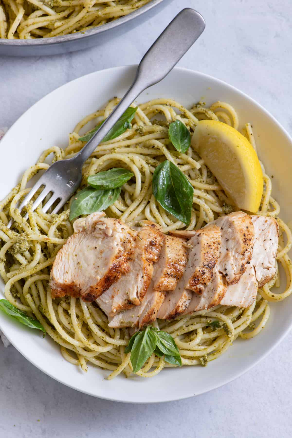 Shallow bowl with the pasta topped with chicken and garnished with lemon and basil with a fork in bowl.