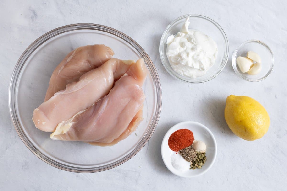 Ingredients for marinated chicken before being prepper with greek yogurt, garlic cloves, whole lemon, spices, and uncooked chicken breast in bowl.