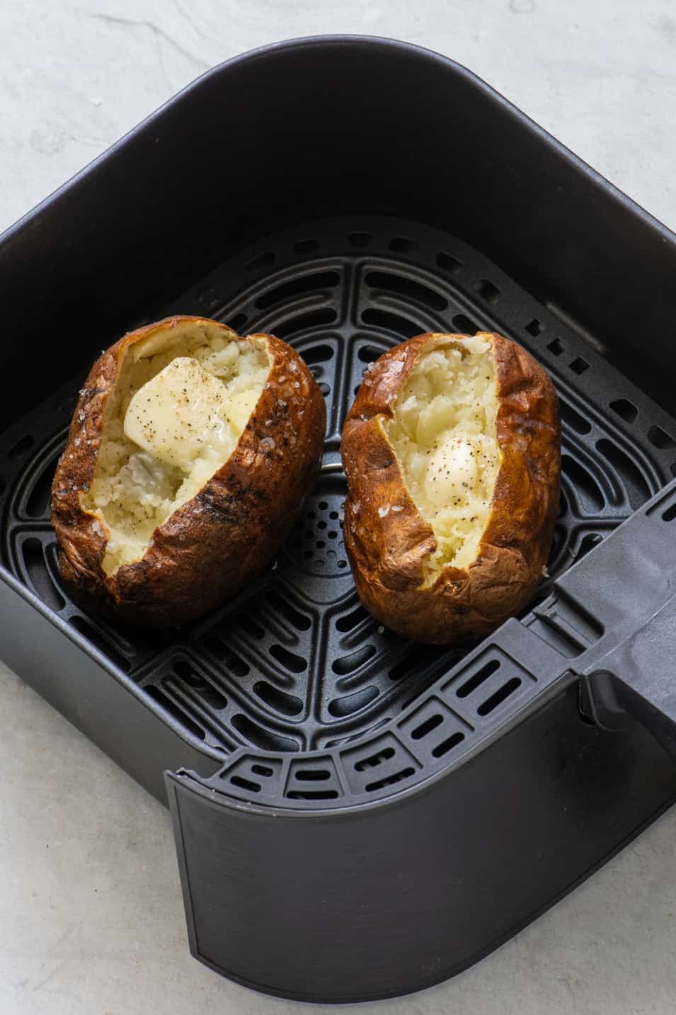 2 baked potatoes in an air fryer basket split open with butter, salt, and pepper.