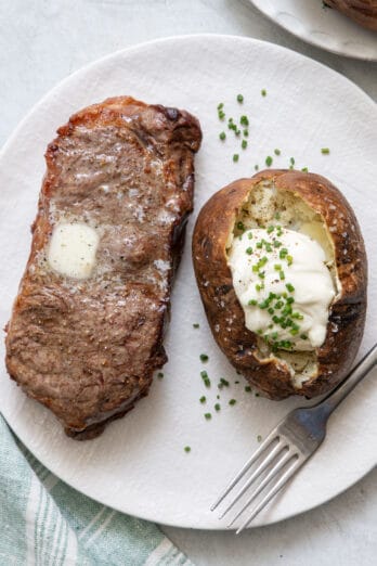 Air fryer steak on a textured plate with melted butter on top next to baked potato topped with sour cream and chives.